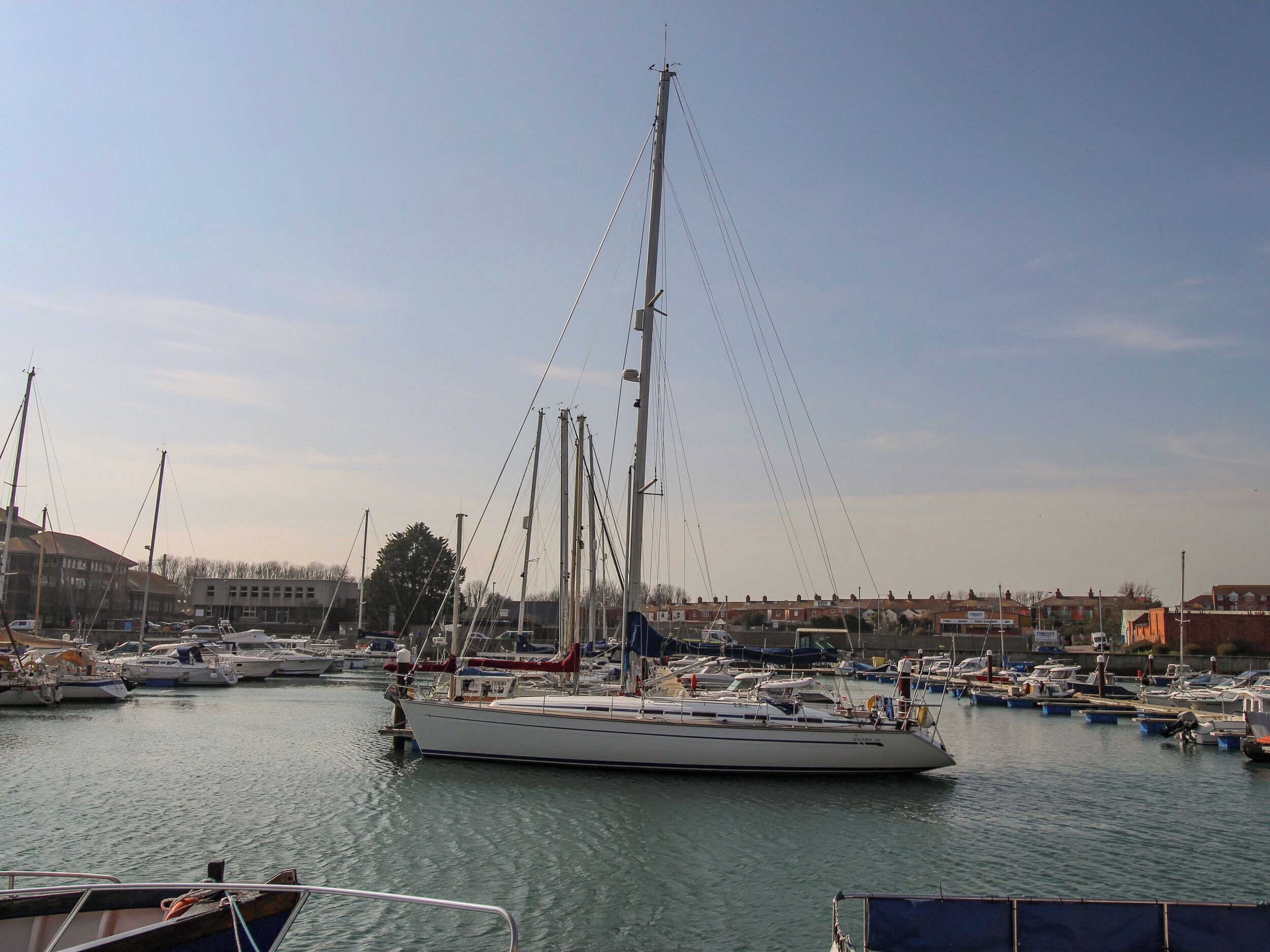 A marina with multiple boats docked, a large sailboat in the foreground, and buildings in the background.