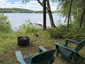 A lakeside view with a dock, a fire pit, and two green Adirondack chairs.