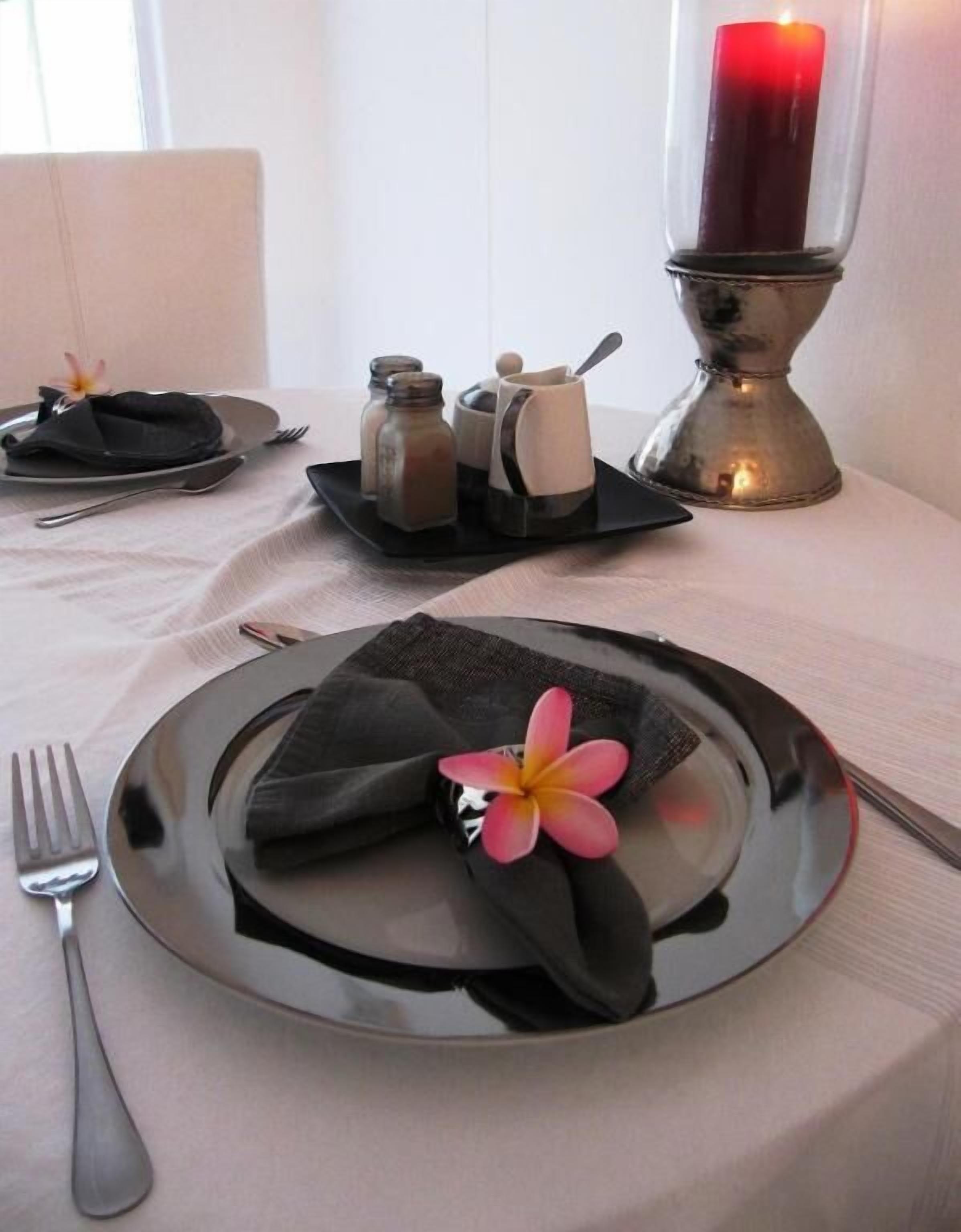Una mesa dispuesta con un plato, servilleta y una flor. Hay saleros y pimenteros, una vela, además de un juego de salero y pimentero sobre la mesa.
