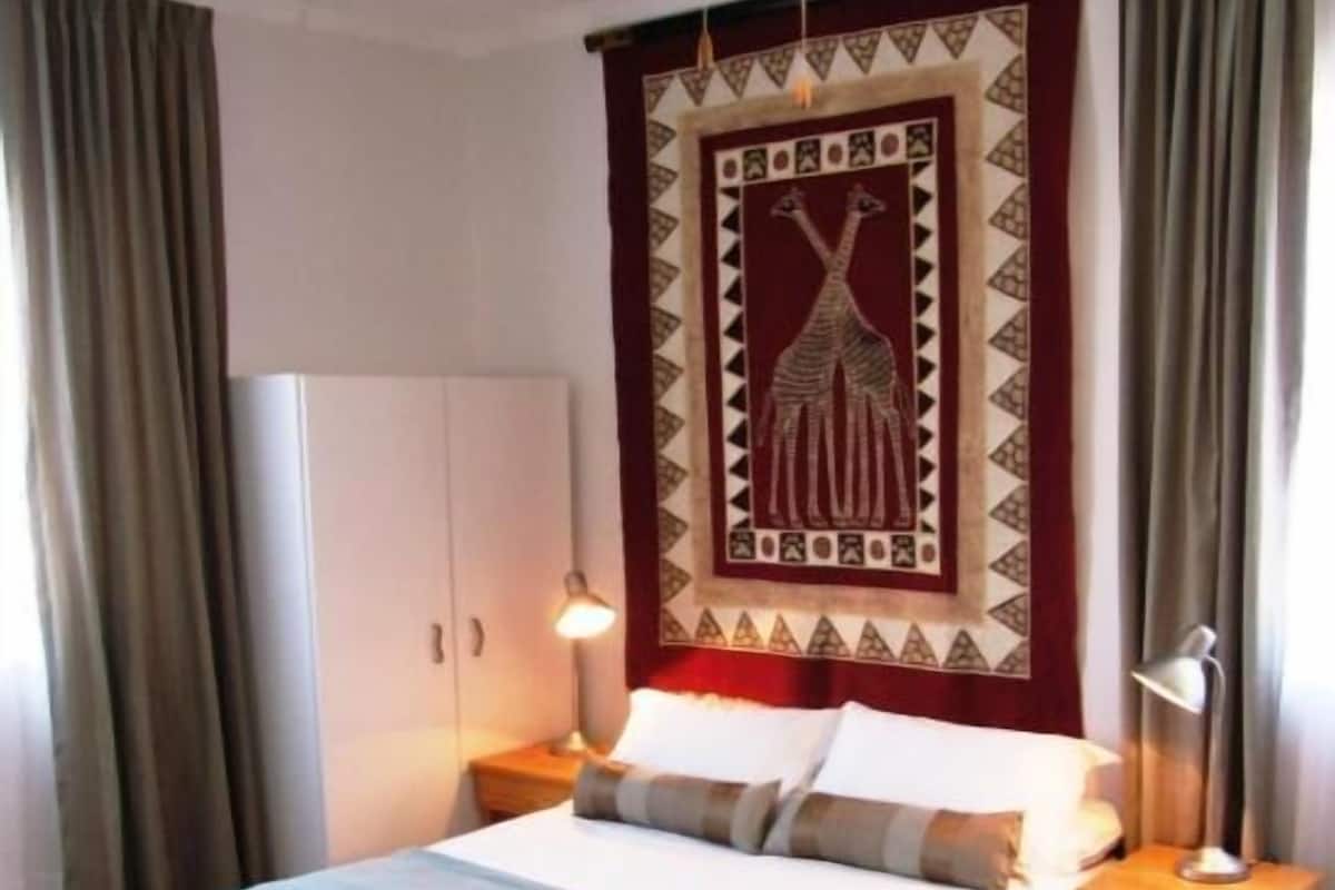 A bedroom with a bed, a ceiling fan, a nightstand with a lamp, and a wall tapestry.