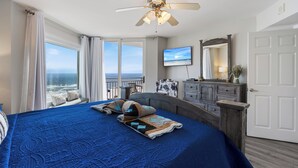 Un dormitorio con una cama grande, ventilador de techo, televisor y vista al océano.