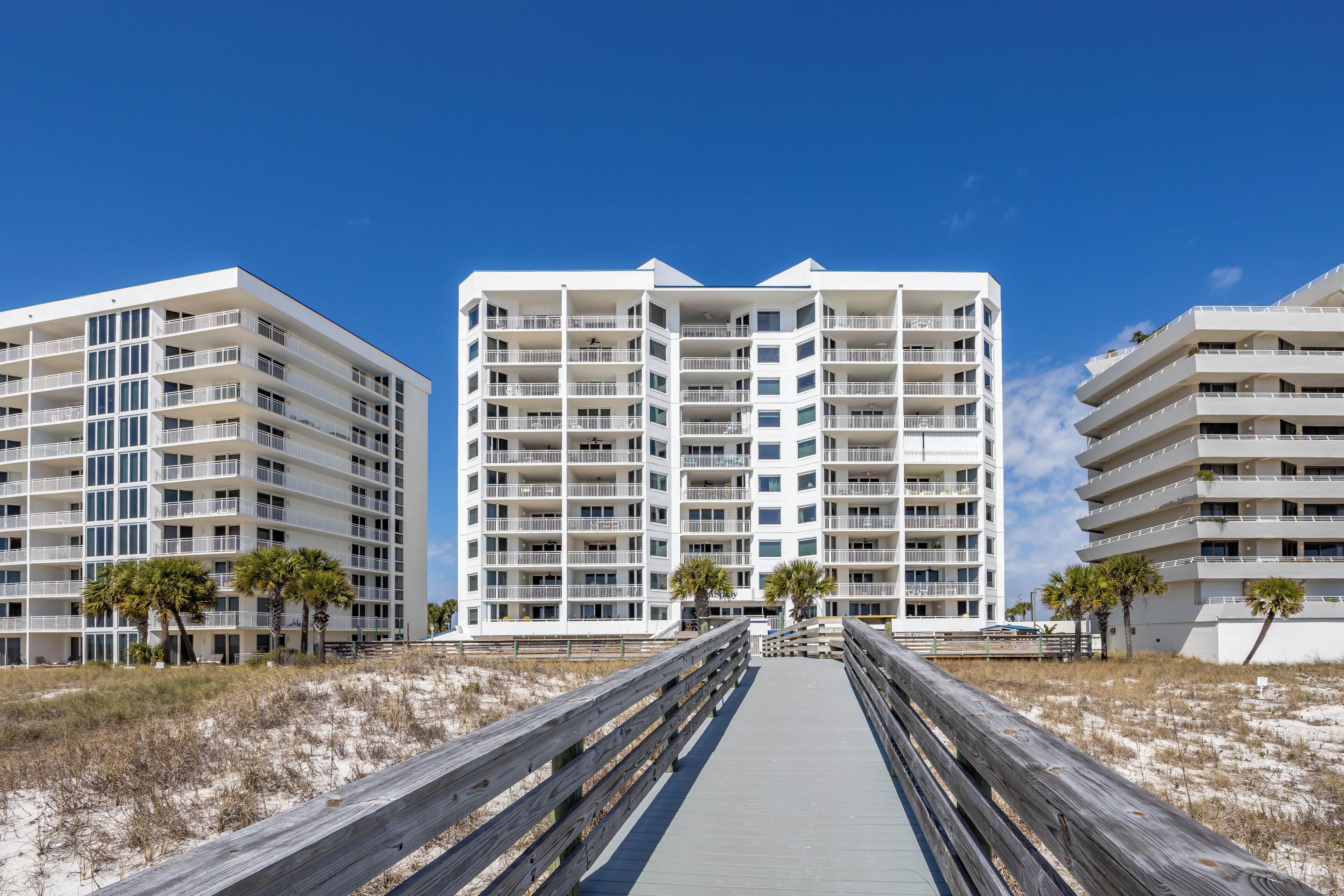 A beachfront hotel with multiple balconies, a boardwalk leading to the entrance, and palm trees.