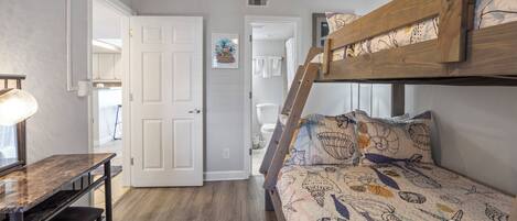 A bedroom with a bunk bed, a desk with a lamp, and a door leading to a bathroom.