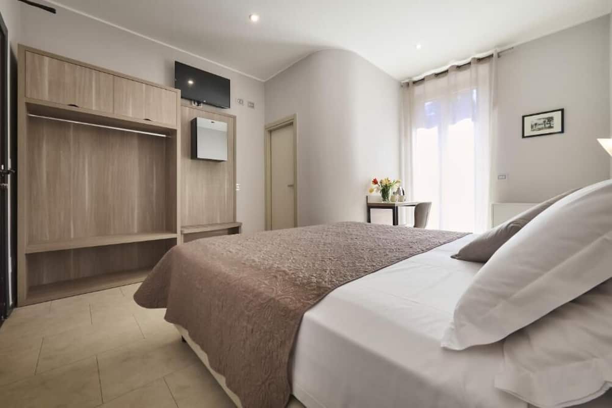 A modern hotel room with a large bed, a wardrobe, a flat-screen TV, and a small table with a vase of flowers.