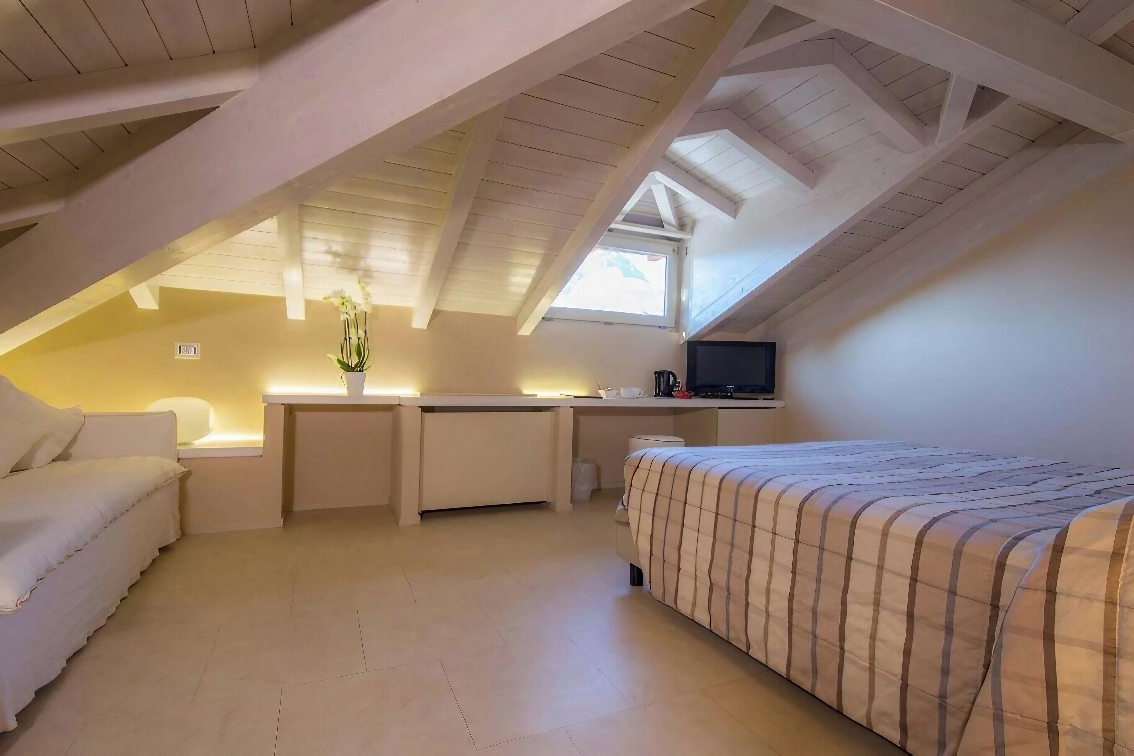A bedroom with a sloped ceiling, a bed with a striped bedspread, a desk with a chair, a small window, and a TV on the wall.