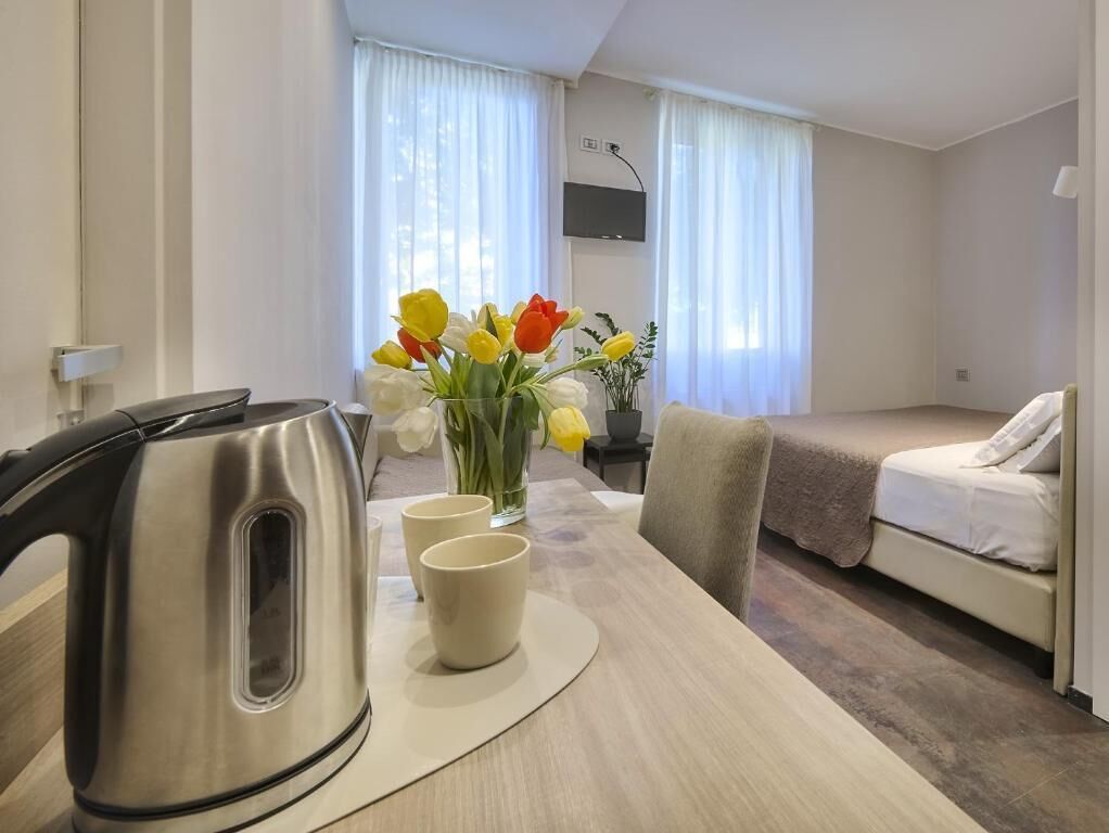 A hotel room with a bed, a table with a kettle and cups, a vase with flowers, and a TV on the wall.