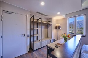 A modern room with a built-in desk, a chair, a lamp, a window with a mountain view, and a wardrobe with shelves.