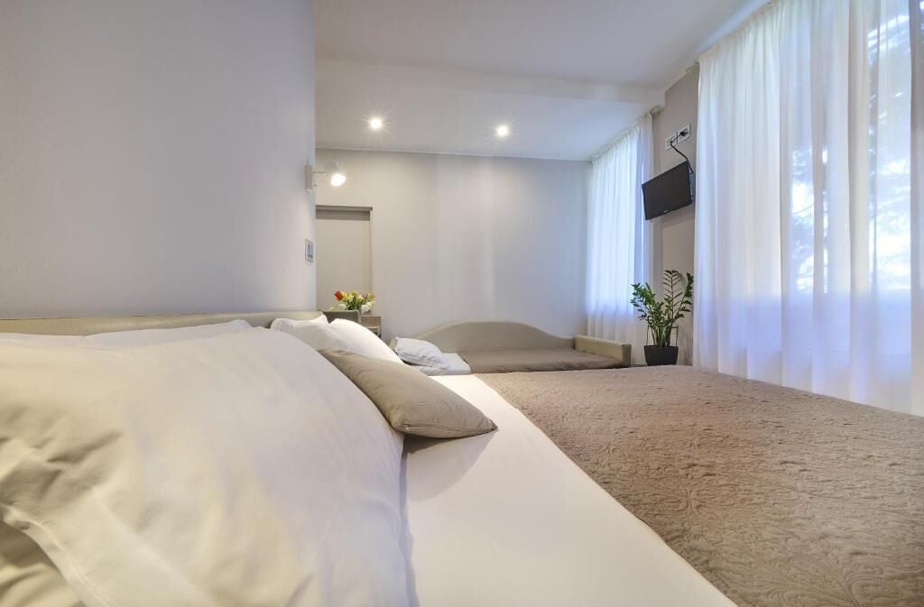 A hotel room with two beds, a flat-screen TV mounted on the wall, a potted plant, and a large window with sheer curtains.