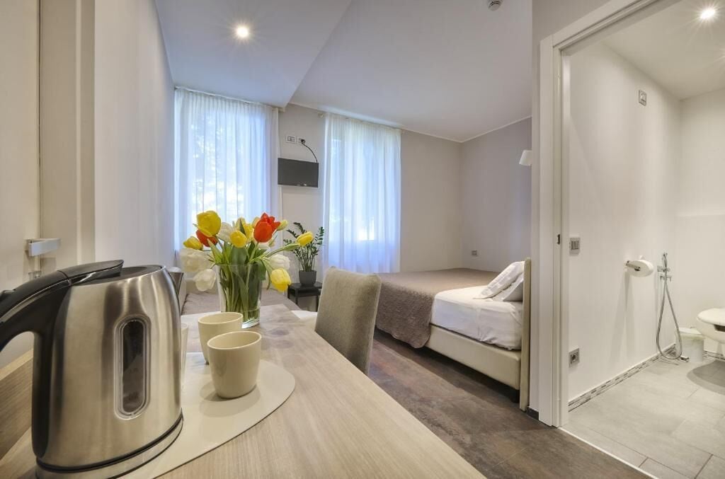 A compact hotel room with a bed, a small table with a kettle and cups, a vase with flowers, and a shower area.