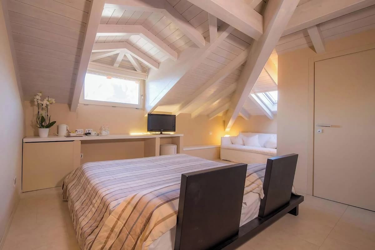 A bedroom with a sloped ceiling, a bed with a striped bedspread, a desk with a television, and a window providing natural light.