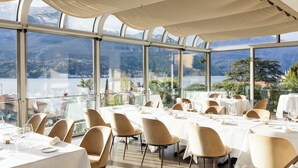 Restaurante com mesas preparadas para refeições, grandes janelas e vista para montanhas e água.