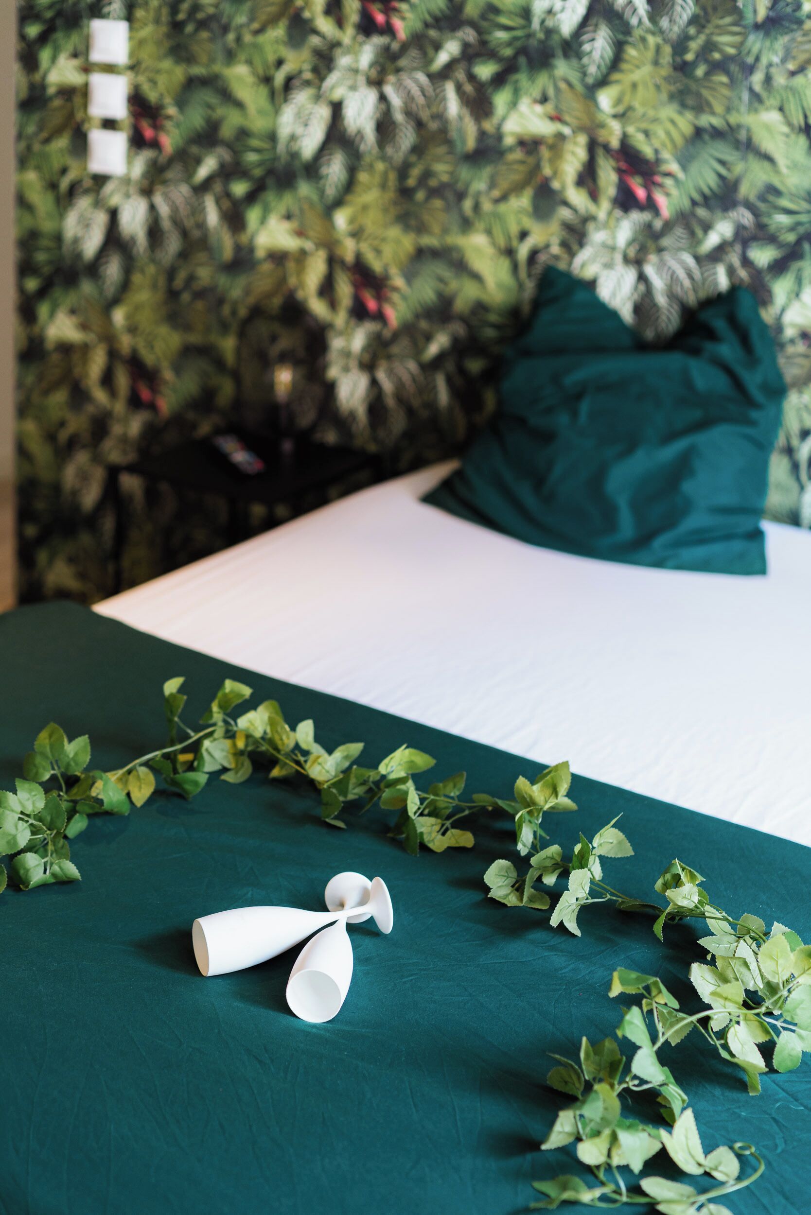 Un llit ben fet amb un estovat verd i coixins blancs, una enfiladera decorativa amb flors blanques, i un fons de paper pintat amb dibuixos.