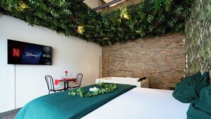 A hotel room with a bed, a TV displaying streaming services, a brick wall, and a green plant wall.