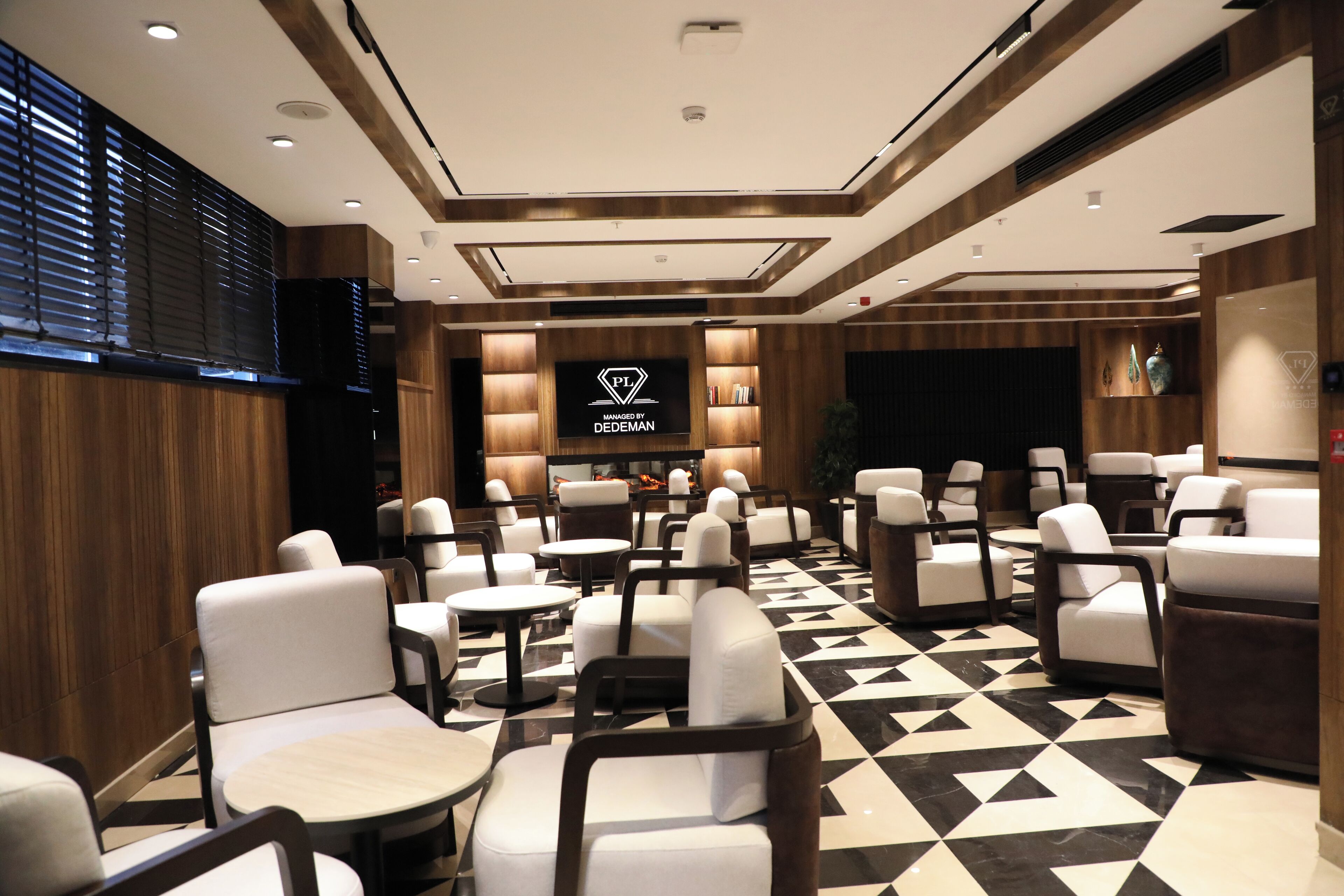 A hotel lobby with a checkered floor, multiple seating areas, and a central TV screen displaying the logo "PL Managed by DEDEMAN".