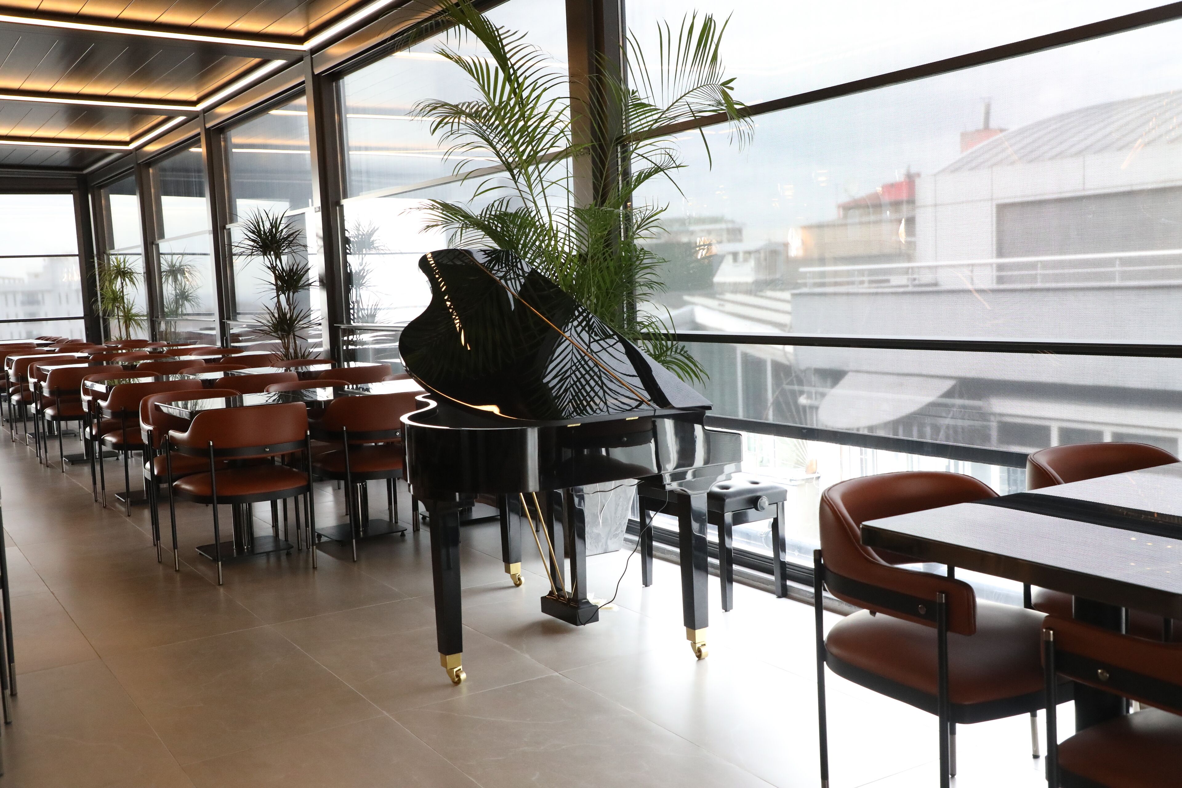 A spacious restaurant with a piano, red chairs, and large windows offering a city view.