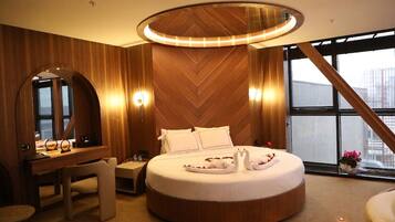 Quarto de hotel moderno com uma cama redonda, painéis de madeira, uma escrivaninha e uma janela grande.
