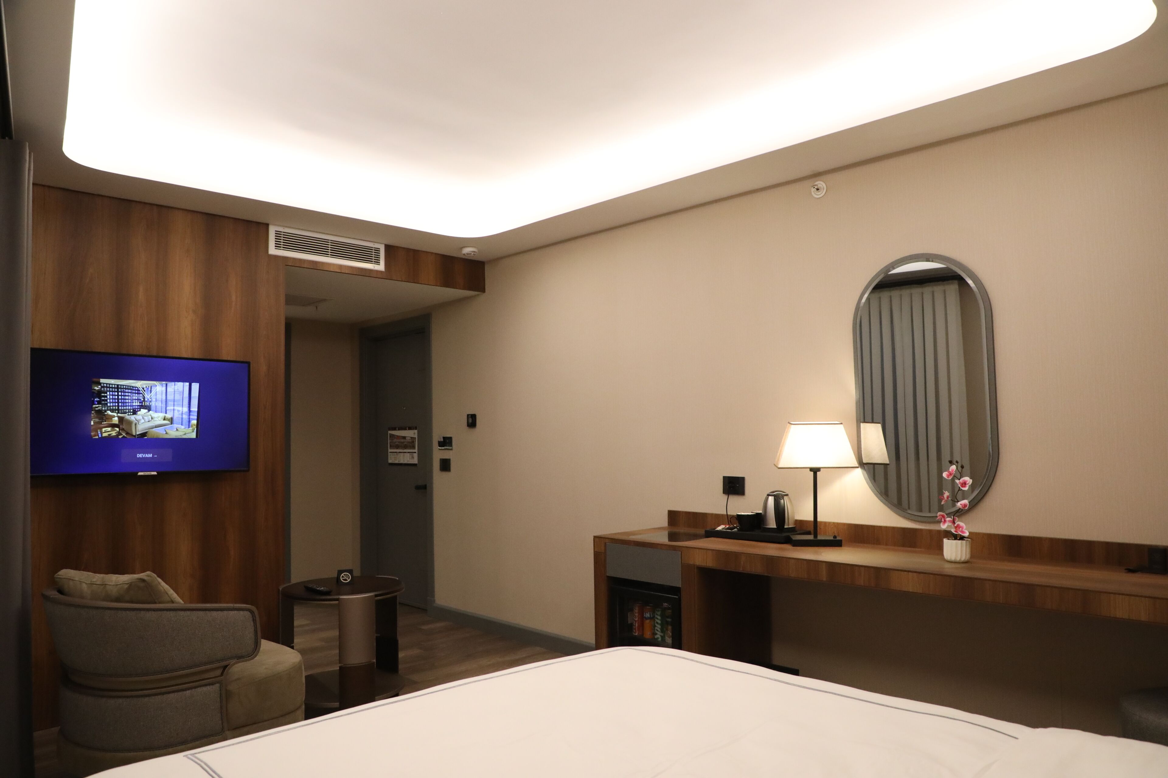 A hotel room with a bed, a TV mounted on the wall, a desk with a lamp, a chair, and a small table.