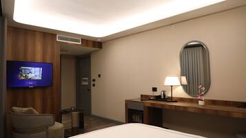 A hotel room with a bed, a TV mounted on the wall, a desk with a lamp, a chair, and a small table.