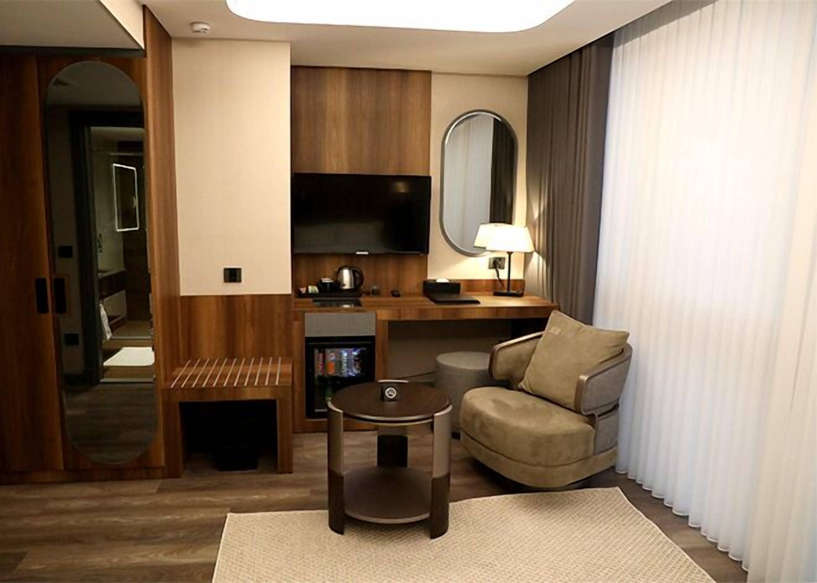 Quarto de hotel compacto com sofá, uma pequena mesa, televisão, espelho e área de cozinha.