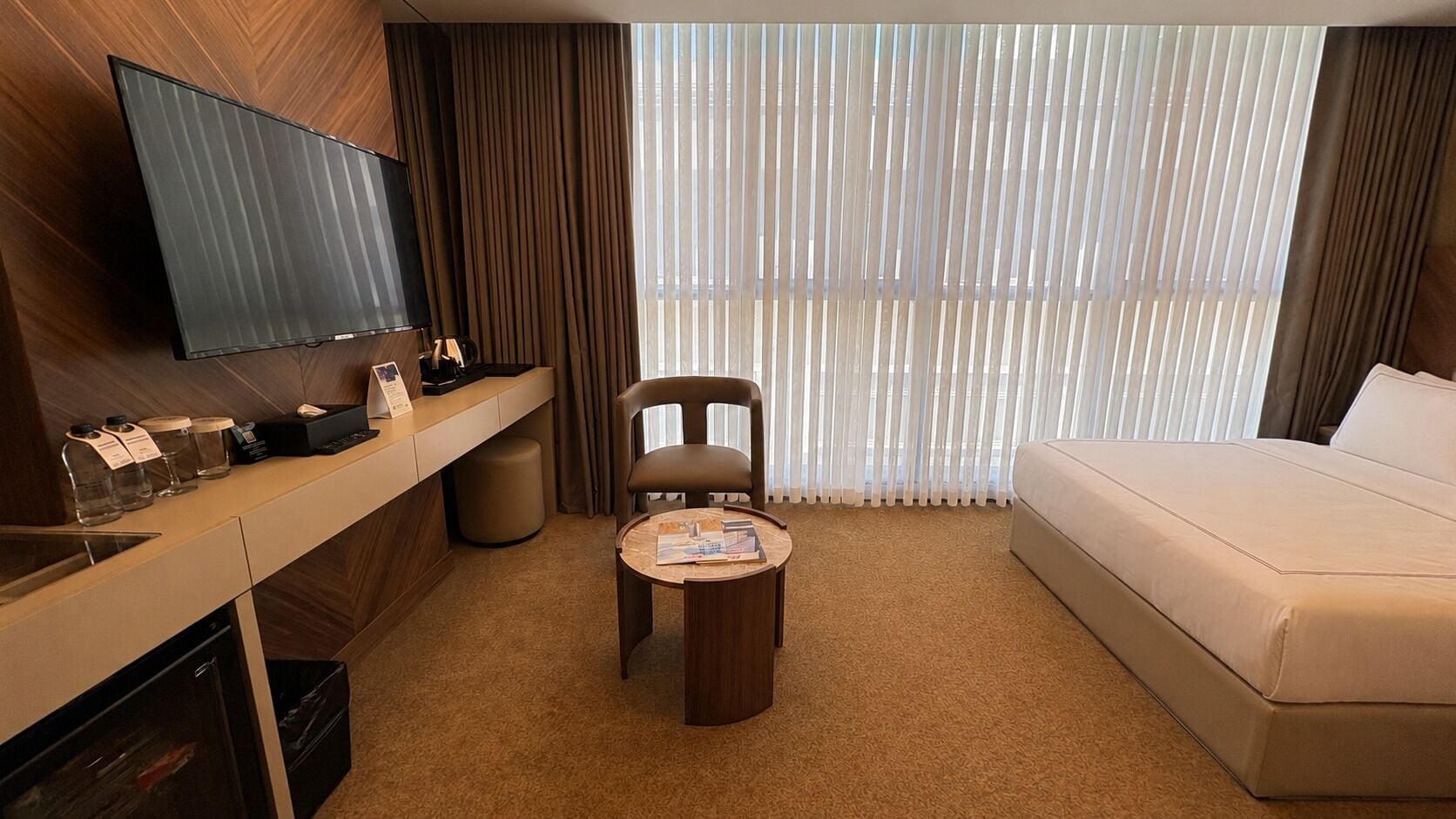 A hotel room with a bed, a desk with a flat-screen TV, a chair, a small table, and a window with blinds.