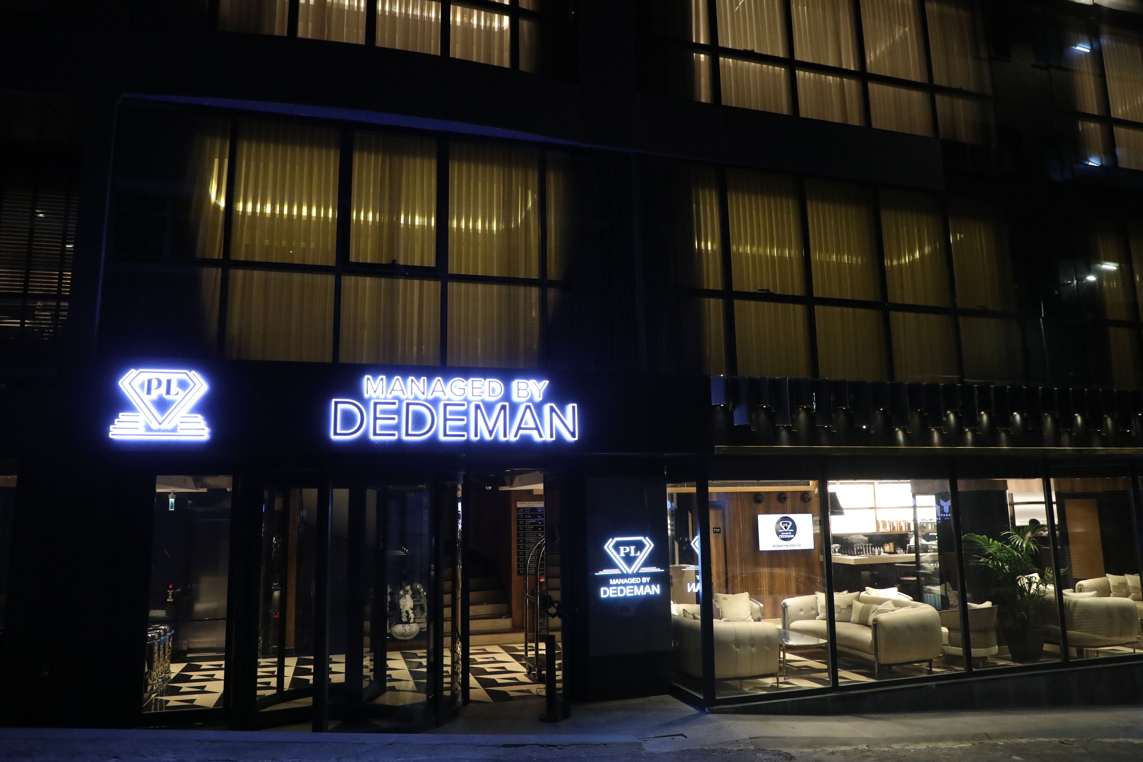 A hotel entrance with a neon sign 'PL Managed by DEDEMAN' and a glass facade displaying furniture.