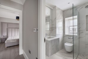A modern bathroom with a glass shower enclosure, a marble-tiled shower area, and a white toilet. - Collingham Apartments (London)