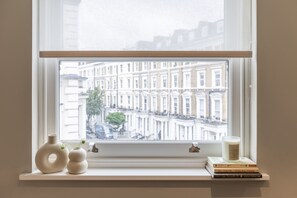 A window with a view of a row of townhouses, a shelf with books and a candle, and a vase with a plant. - Collingham Apartments (London)