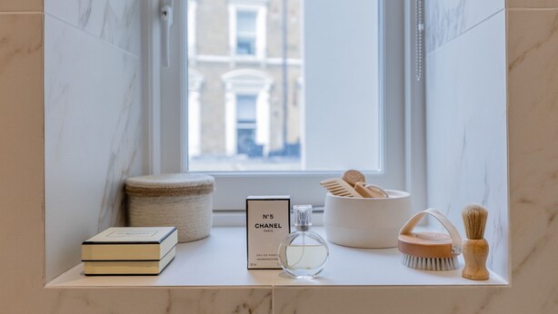 Ein Badezimmer-Waschtisch mit Marmor-Spritzschutz, ein Fenster mit Blick auf Gebäude, eine Chanel-Parfümflasche, ein Buch, eine Holzbürste und ein Holzkästchen.
