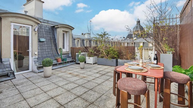 Un patio con una mesa dispuesta para dos personas, rodeado de plantas en macetas y una valla de madera.