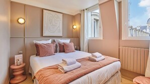 Habitación de hotel con una cama grande, una mesita de noche con lámpara, un cuadro en la pared y una ventana con vistas a edificios.