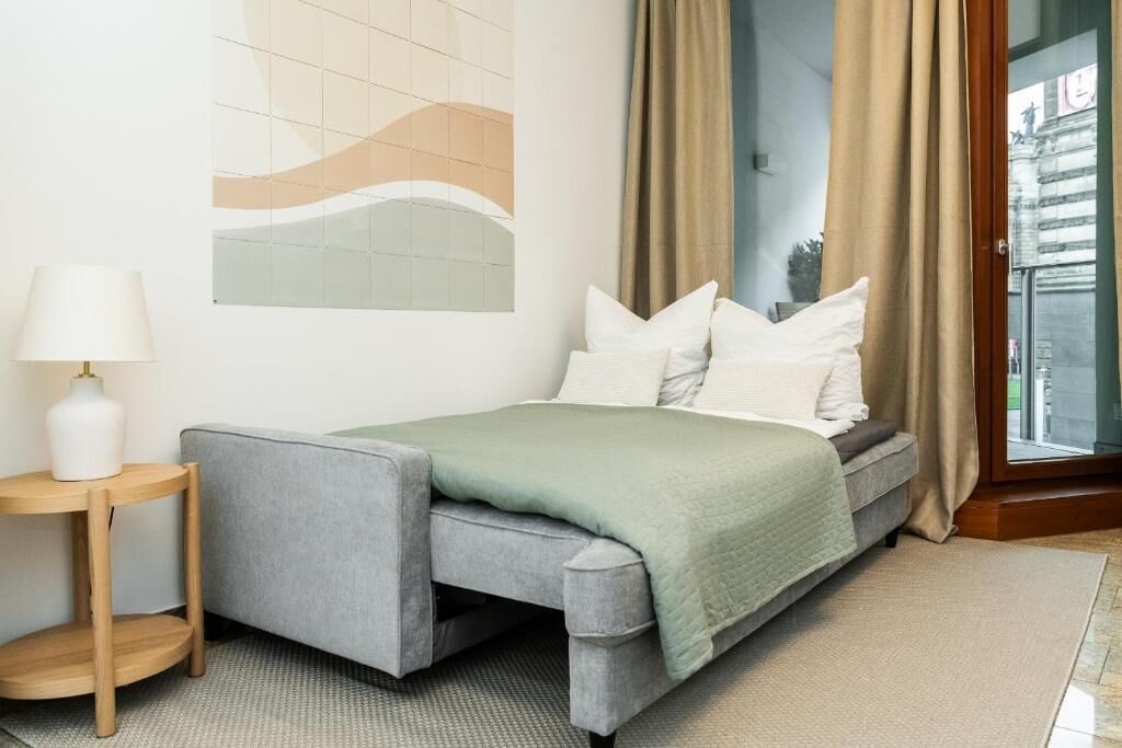 Quarto moderno com um sofá-cama cinza, uma pequena mesa de cabeceira de madeira com um vaso branco, um cobertor verde e uma janela grande com cortinas bege.