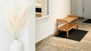 Un couloir avec un banc en bois, un vase blanc contenant des plumes d’herbe des pampas et un tableau encadré accroché au mur.