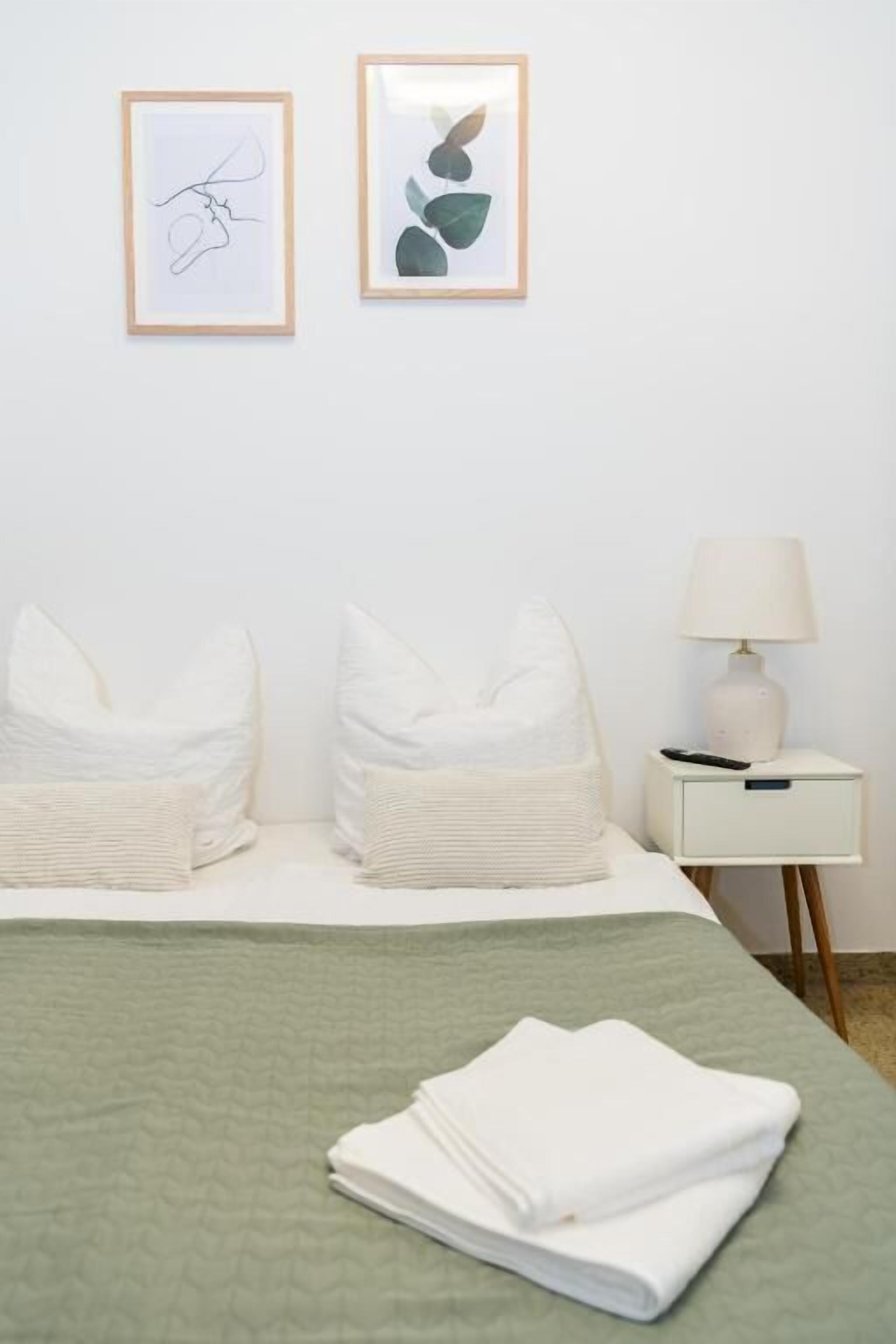 A hotel room with a bed, pillows, a bedside table with a lamp, and two framed artworks on the wall.