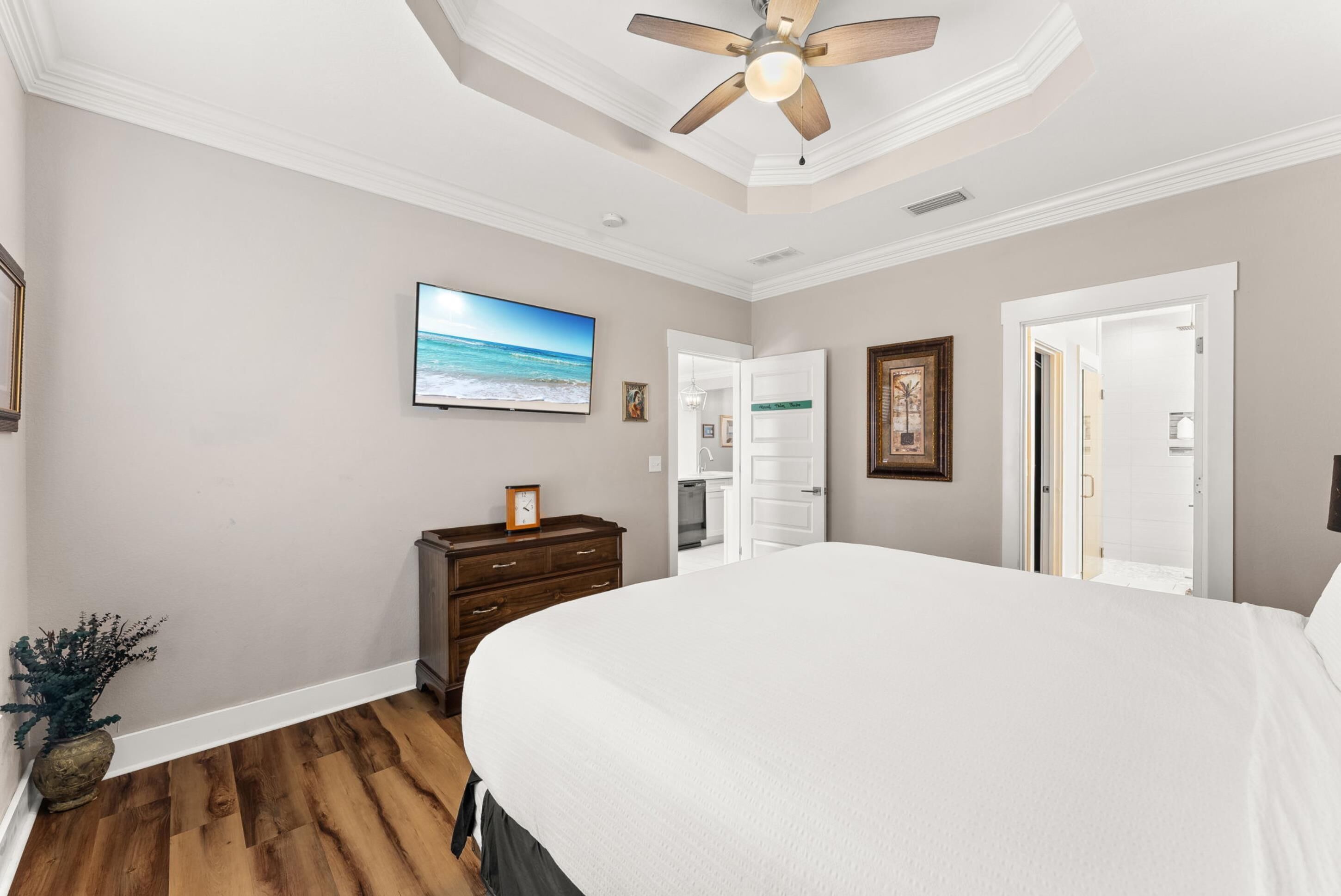 A bedroom with a large bed, wooden flooring, a ceiling fan, a TV on the wall, and a door leading to a bathroom.