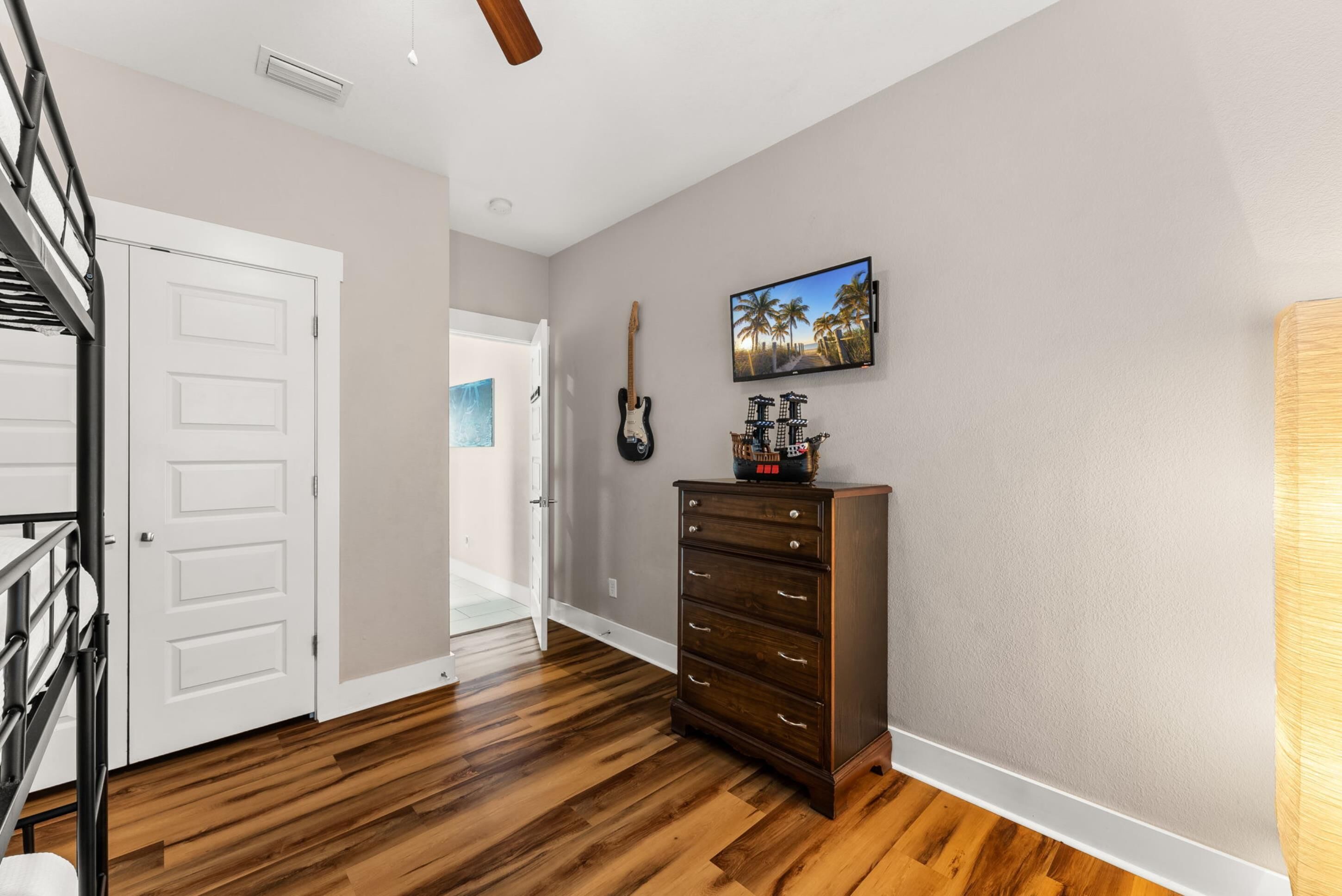 A hallway with wooden flooring, a staircase, a white door, a dark wooden dresser, and a mounted television displaying a beach scene.