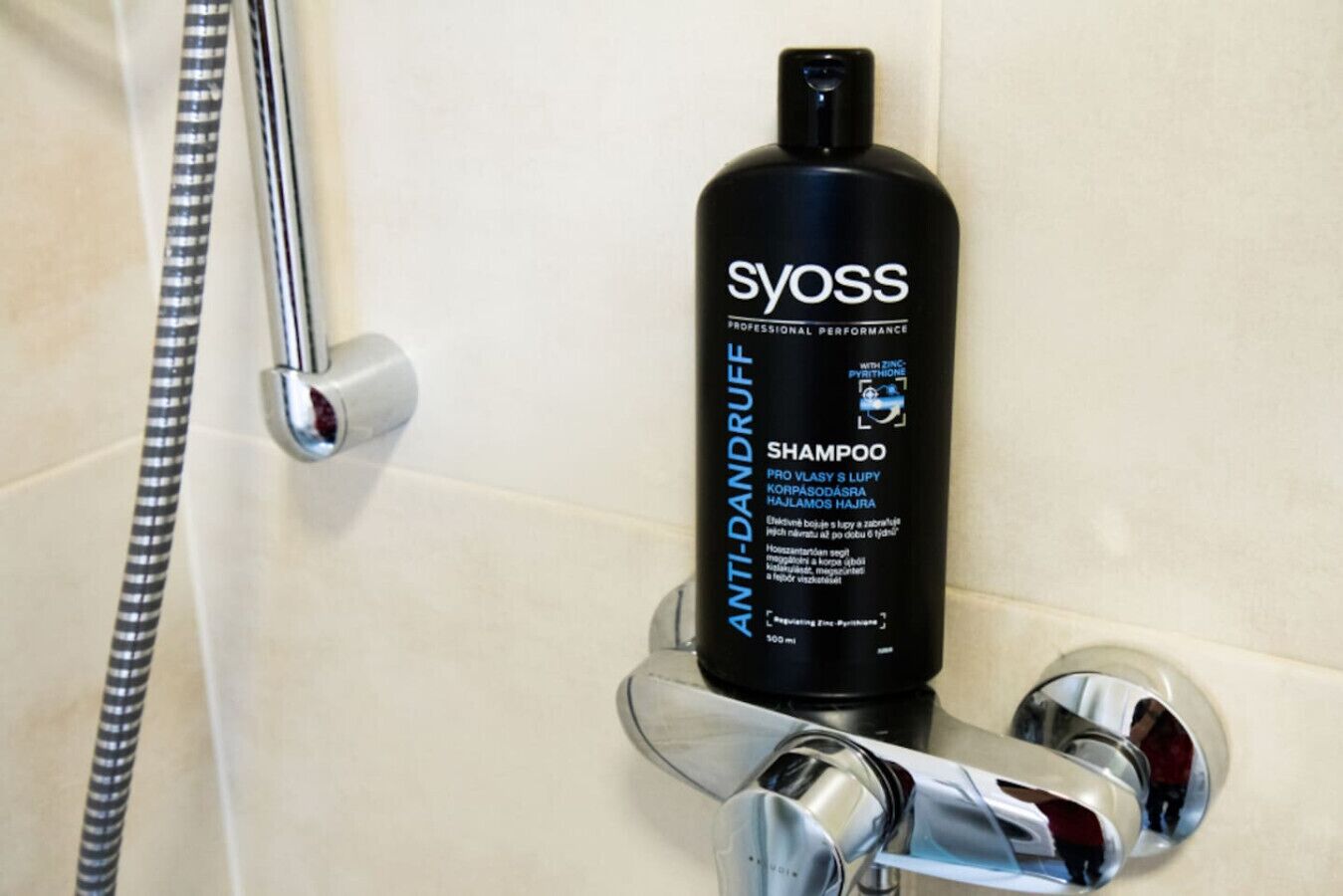 A black bottle of Syoss Anti-Dandruff Shampoo on a bathroom sink with chrome fixtures.