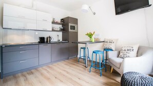 Een moderne keuken met grijze kastjes, een magnetron en een koffieapparaat. Er is een eethoek met een tafel en blauwe krukken. Een flatscreen televisie hangt aan de muur.