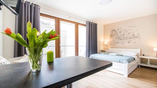 Een moderne hotelkamer met een bed, een bureau met een bloevas, een kaart aan de muur en een balkon met gordijnen.