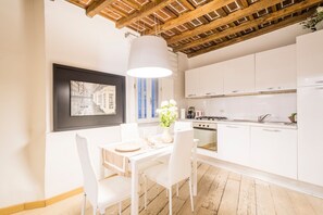 Une cuisine moderne avec des meubles blancs, une table à manger avec des chaises, un luminaire suspendu et un tableau encadré au mur.
