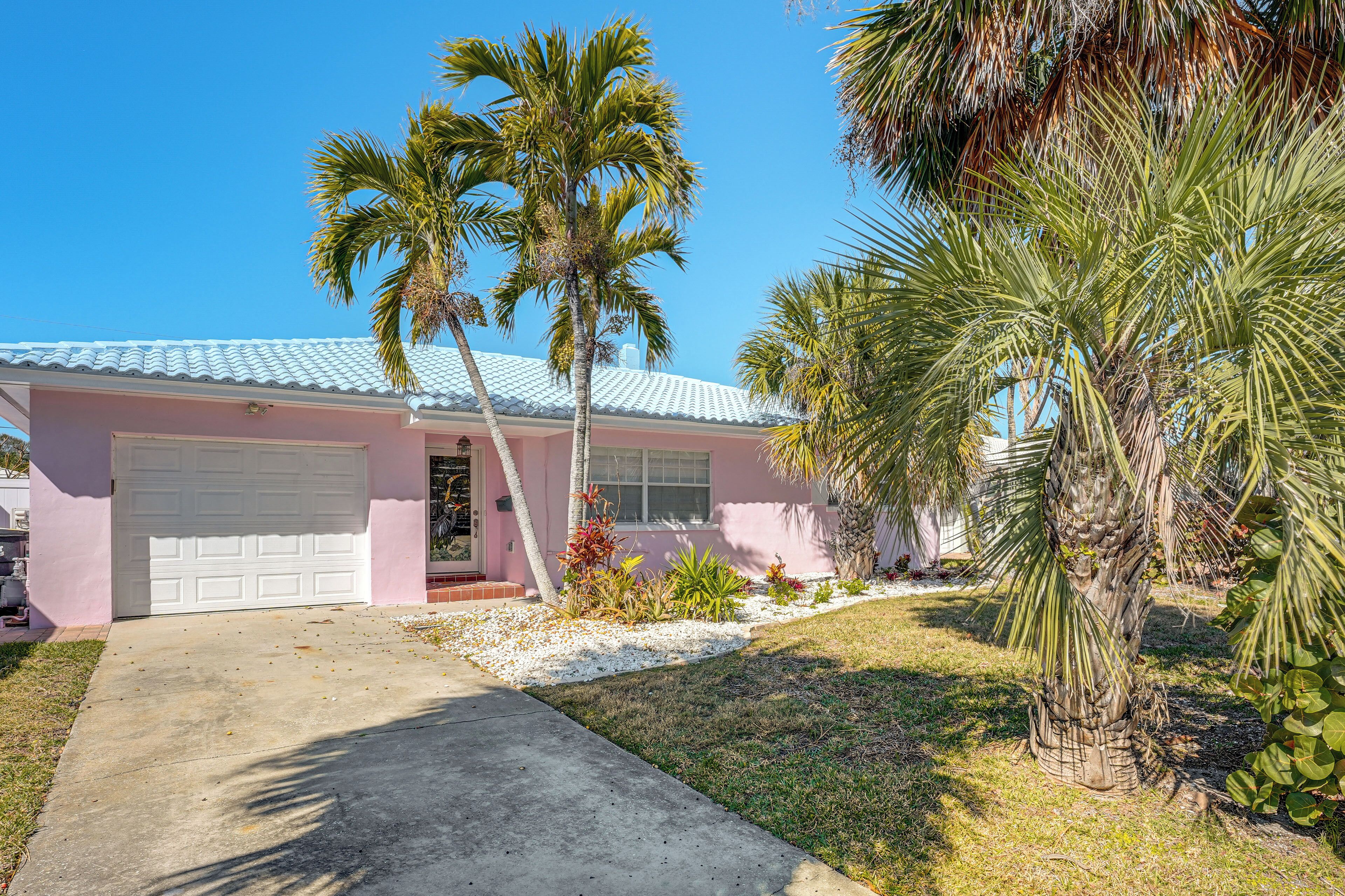 A pink house with a white garage, surrounded by palm trees and a small garden.