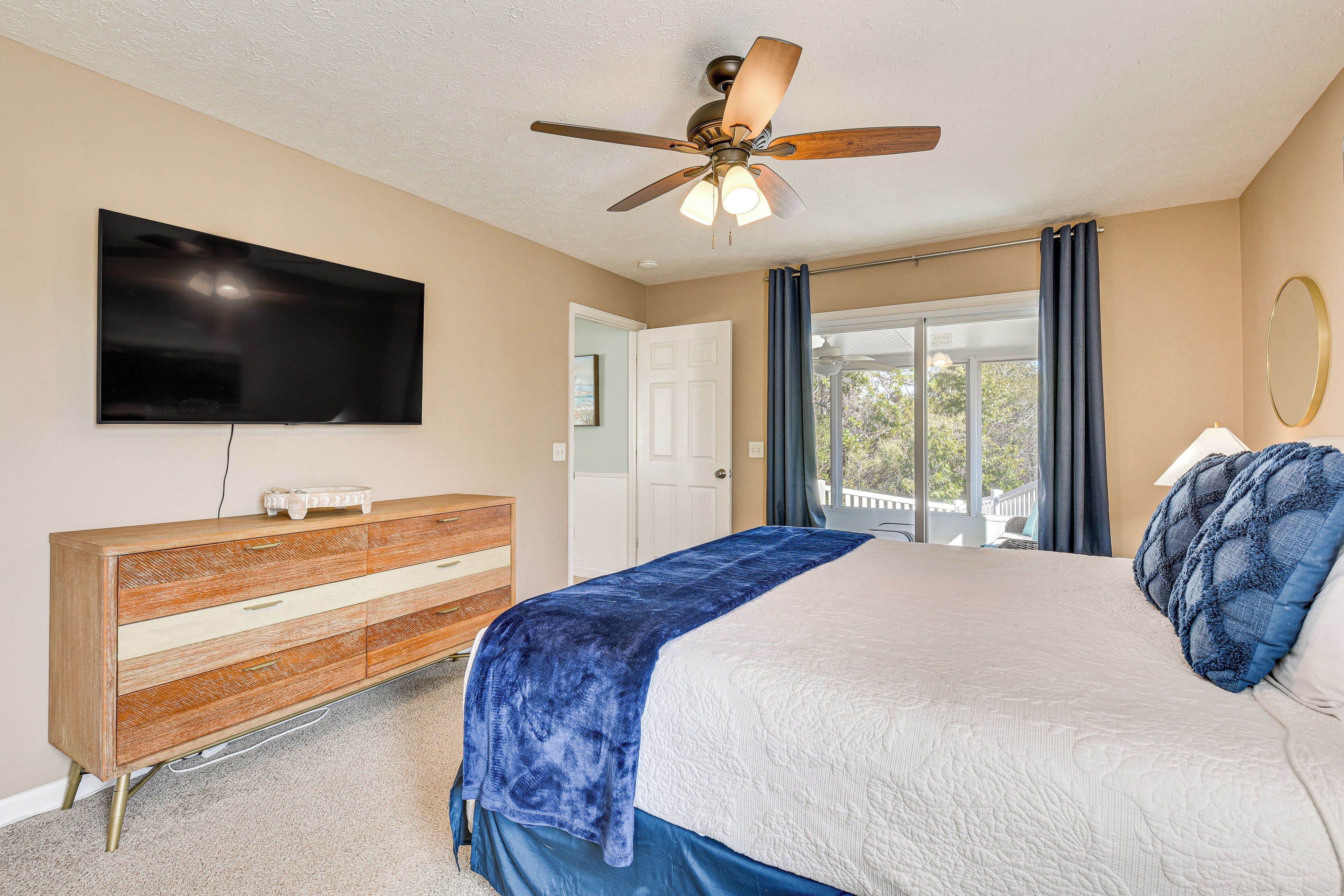 A bedroom with a bed, a ceiling fan, a TV, a dresser, and a view of the outdoors.