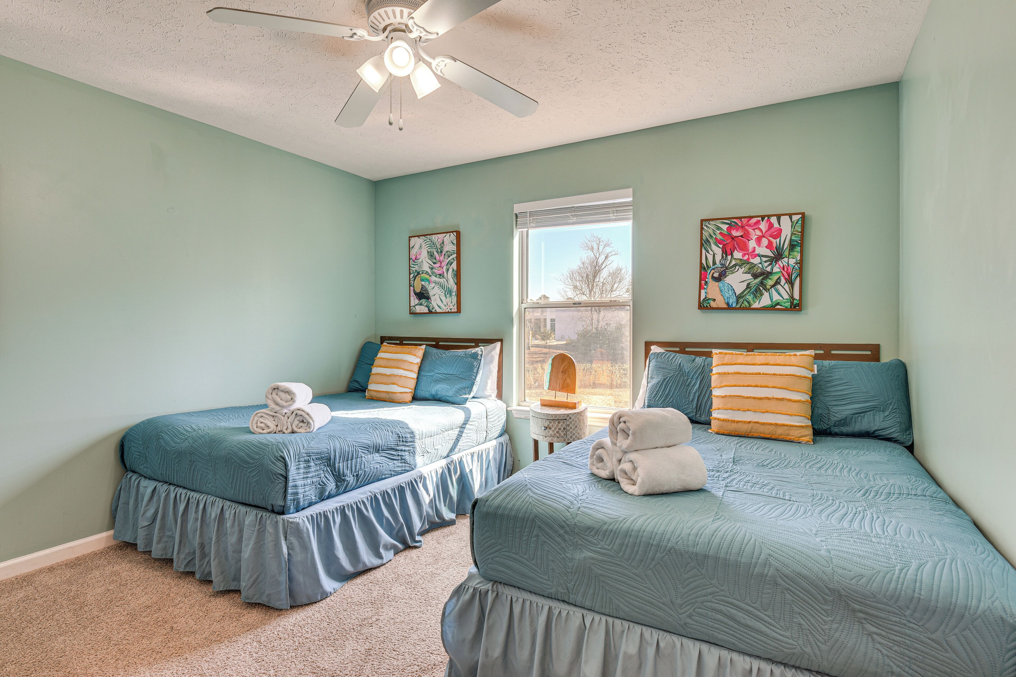 A bedroom with two beds, each with blue bedding and pillows, a ceiling fan, and two wall-mounted paintings.