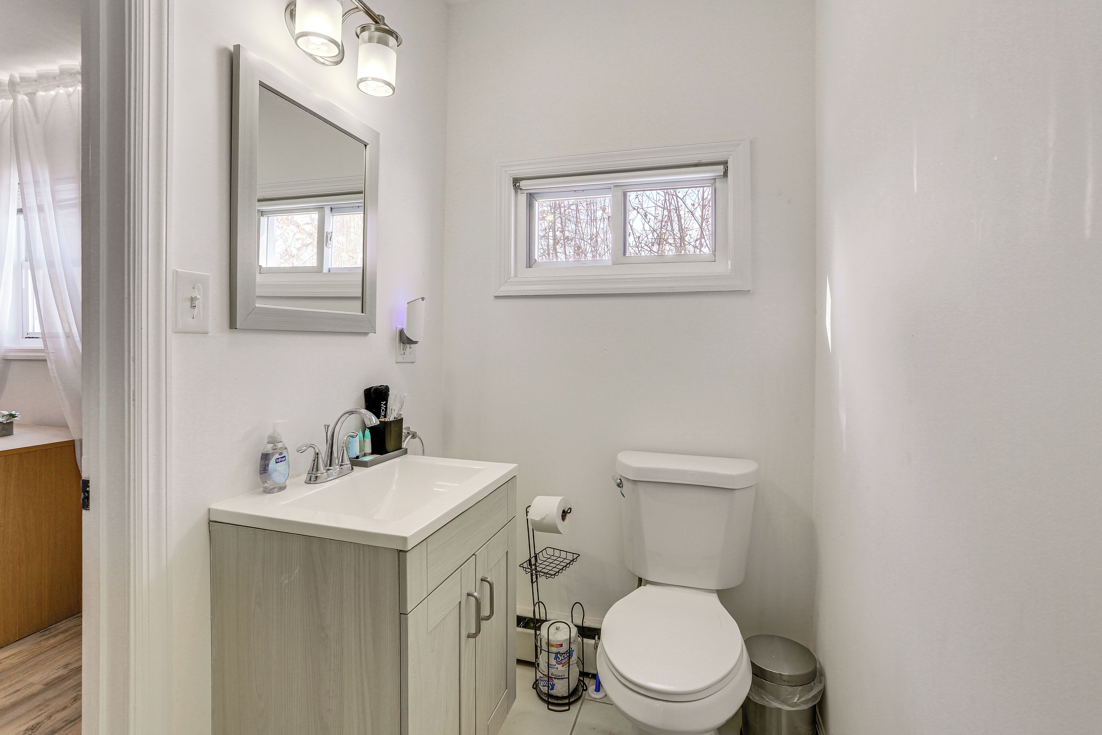 Ein modernes Badezimmer mit einem weißen Waschbecken, einem Spiegel und einer Toilette. Über der Toilette befindet sich ein Fenster und darüber ein kleines Regal mit Pflegeprodukten.