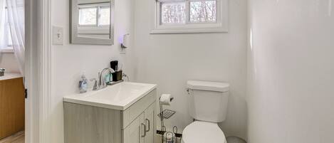 A modern bathroom with a white sink, mirror, and toilet. There is a window above the toilet and a small shelf with toiletries.