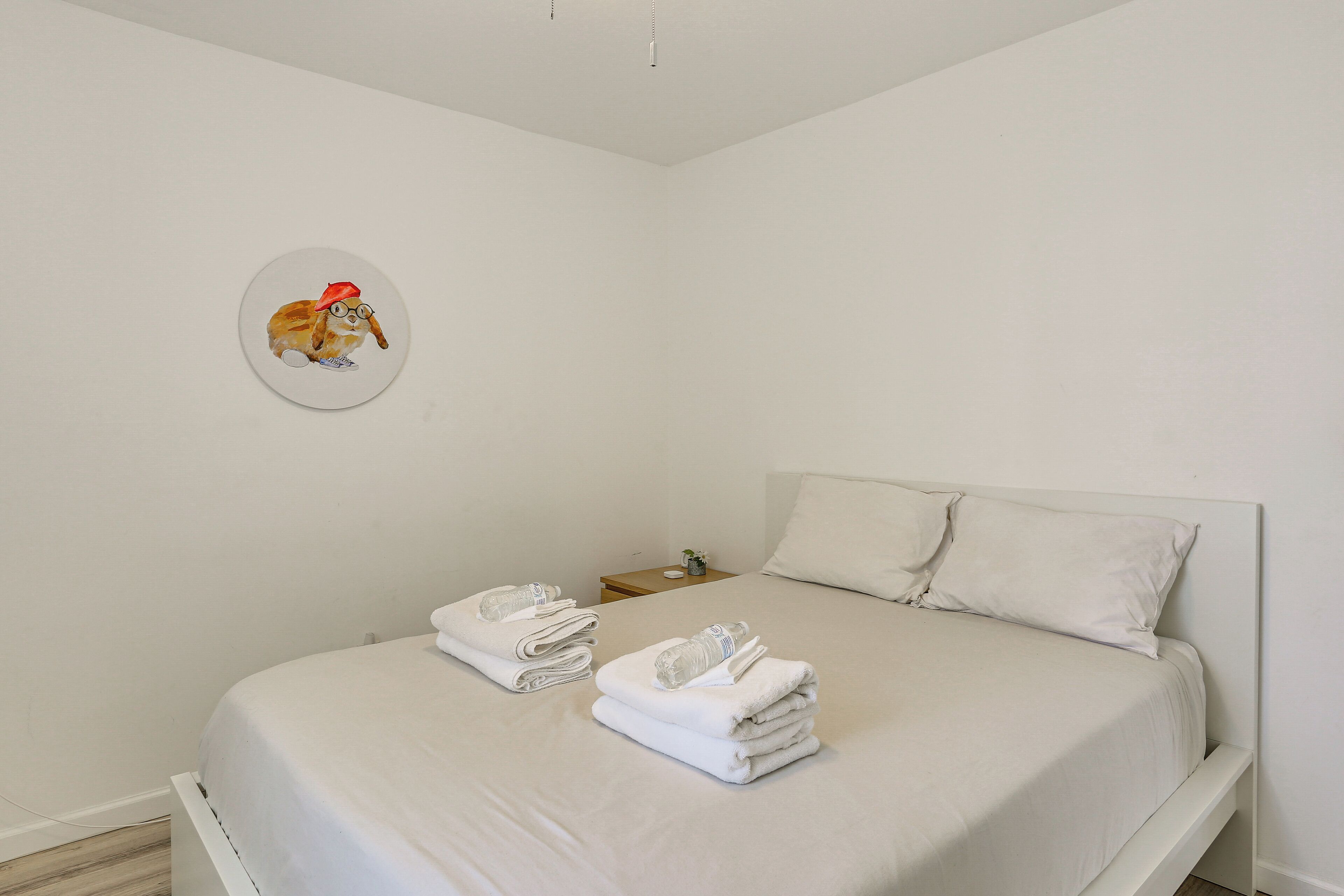 Une chambre à coucher épurée et minimaliste, avec un lit, deux serviettes pliées, une petite table de chevet et un élément décoratif mural.