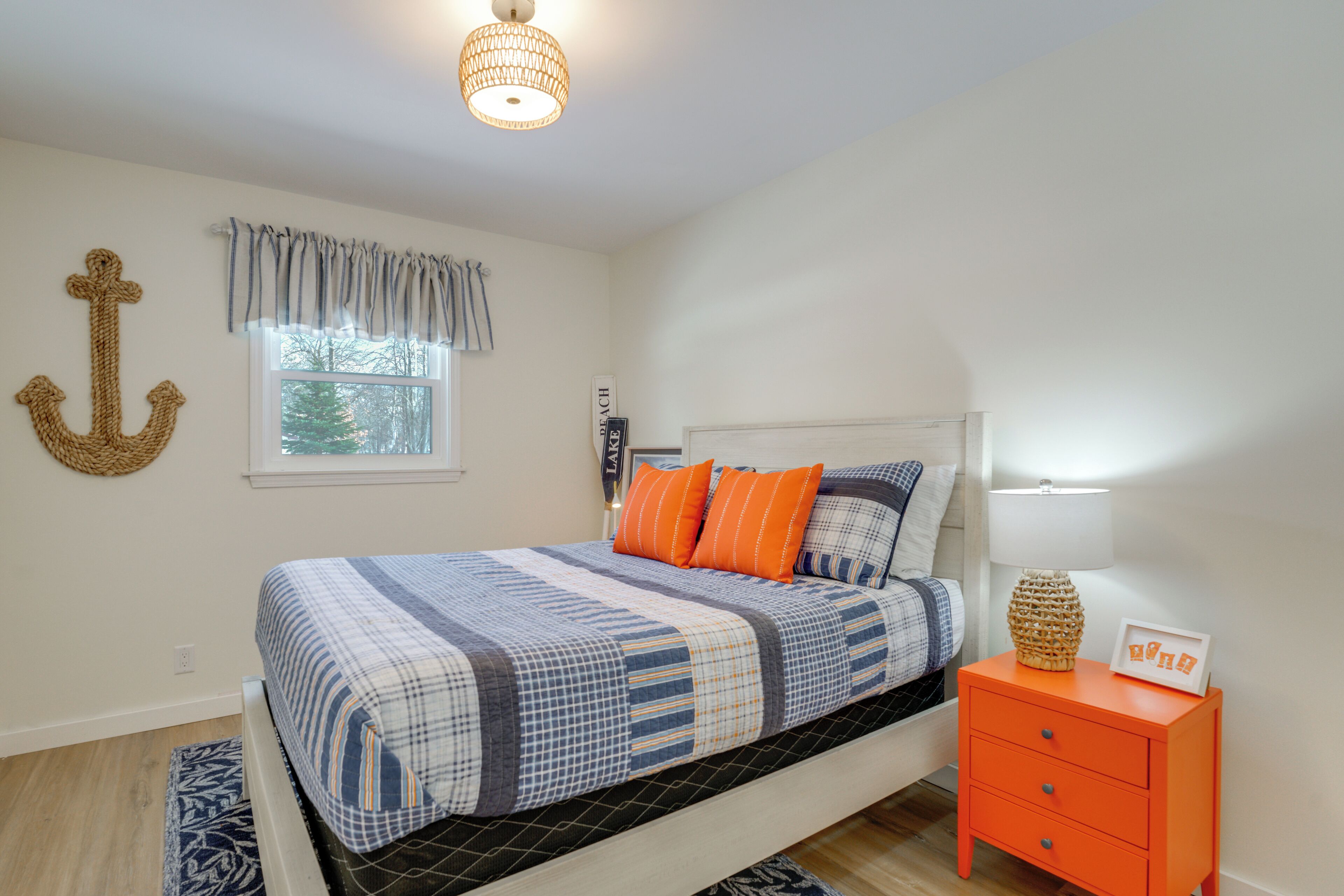 A bedroom with a bed, a nightstand, a lamp, a window, and a wall anchor decoration.