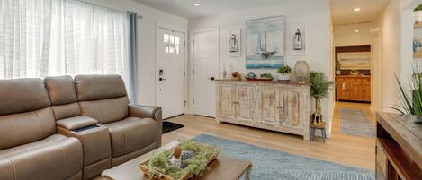 A living room with a brown leather sofa, a wooden coffee table with a decorative item, a blue rug, and a kitchen area with wooden cabinets and a plant.