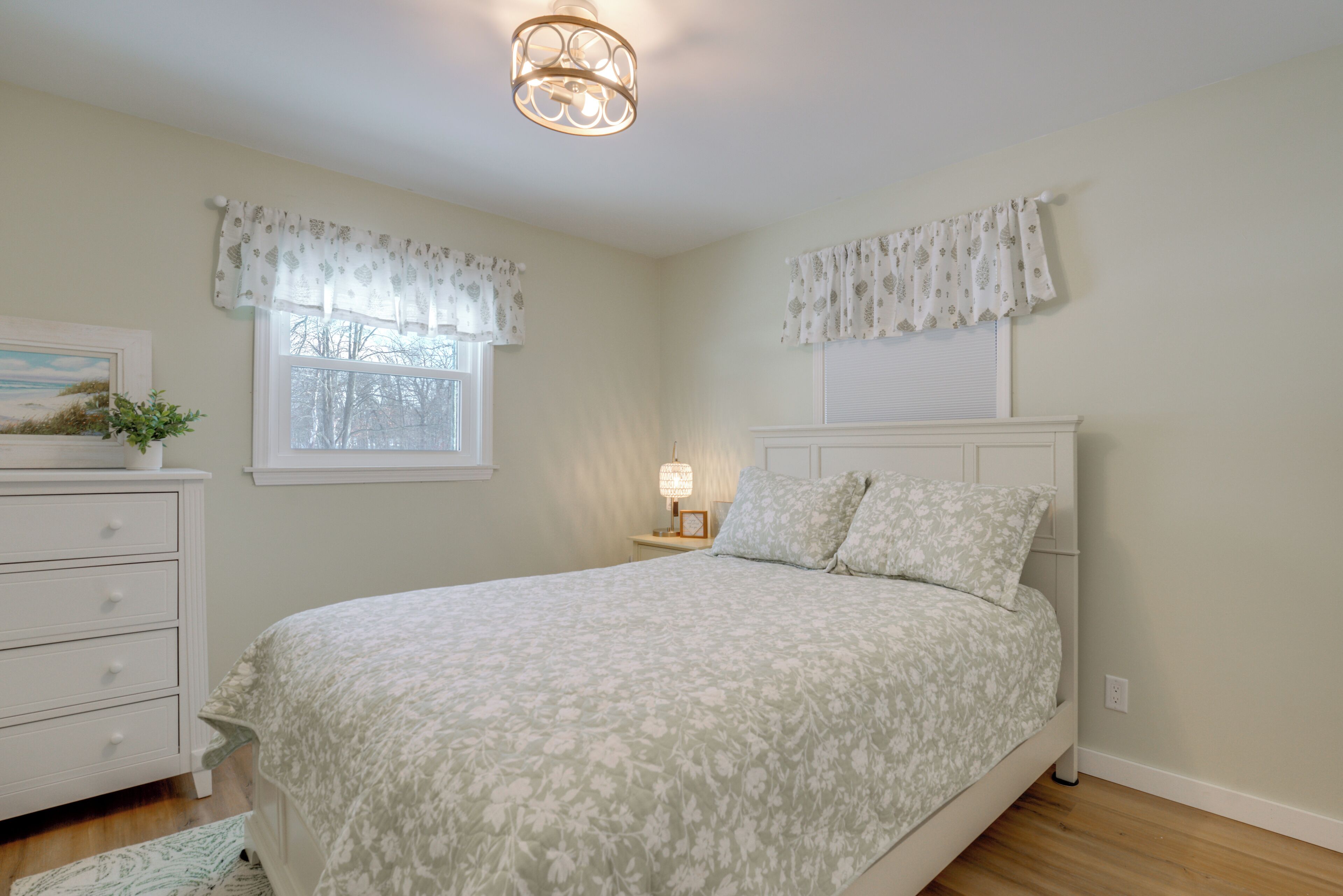 A bedroom with a bed, a nightstand, a lamp, a dresser, and a window with curtains.