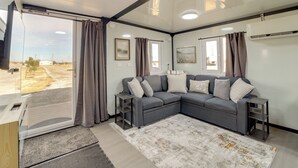 A modern living room with a sectional sofa, a glass door leading outside, and a large window with curtains. - Hunt, Fish & Explore! Tiny Home Near Oologah WMA (Nowata)