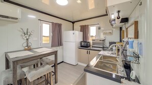 A compact kitchen with a refrigerator, microwave, and sink. There is a wooden table with benches and a small decorative plant. - Hunt, Fish & Explore! Tiny Home Near Oologah WMA (Nowata)